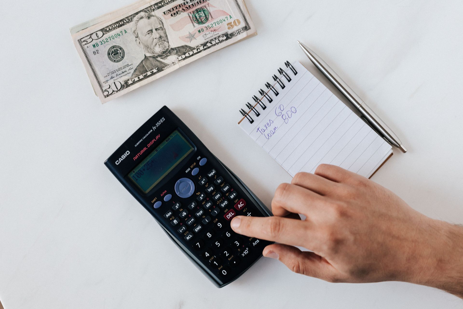 Top view of crop unrecognizable economist calculating on calculator on table with notepad and pen near pile of dollar banknotes on marble table